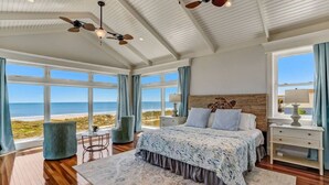 Quarto com uma grande janela com vista para a praia, uma cama com colcha estampada e um ventilador de teto.