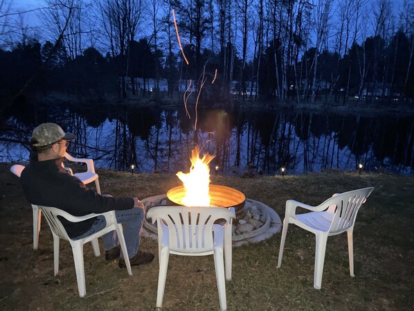 Een persoon zit aan een tafel met een vuurkuil in het midden, omringd door stoelen, buiten in de buurt van een waterpartij.