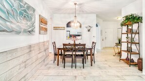 Un comedor con mesa y sillas de madera, una lámpara de araña y un cuadro grande con motivos florales en la pared.