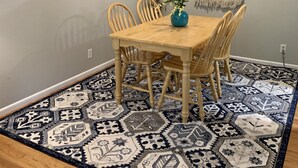 A dining area with a wooden table and chairs, a blue vase with flowers, a patterned rug, and a wall art piece.