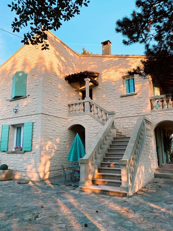 A stone building with a staircase leading to a balcony, a small table with a green tablecloth, and a potted plant.