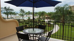 A balcony with a glass table and chairs, a blue umbrella, and a view of a pool and palm trees. - Paradise apartment in beach condo Bay View Grand (Puerto Vallarta)