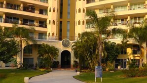 A hotel entrance with a curved driveway, palm trees, and a sign with a clock. - Paradise apartment in beach condo Bay View Grand (Puerto Vallarta)