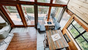 Un interior de madera con ventanas amplias, una mesa de comedor y vistas a una terraza y follaje exterior.