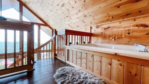 Interior de una cabaña de madera con escalera de madera, un ventanal grande y un baño con lavamanos y ducha.