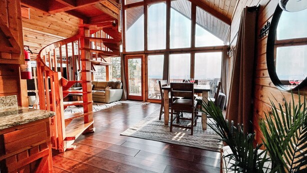 Una habitación amplia con una escalera de madera, un comedor con mesa y sillas, y grandes ventanas que ofrecen una vista.