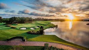 Campo de golf con un cuerpo de agua reflejando el atardecer, rodeado de árboles y un cielo despejado.