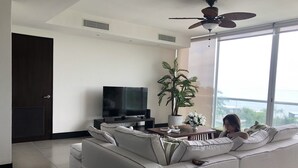 A modern living room with a white sofa, a flat-screen TV, a ceiling fan, a potted plant, and a view of the ocean through large windows.