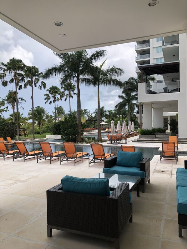 Un espace aménagé au bord de la piscine, avec des chaises longues et un coin salon, entouré de palmiers et bordé d’un bâtiment moderne.
