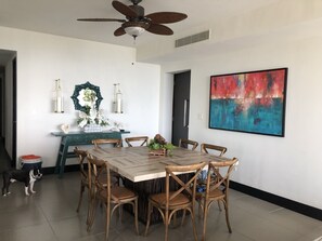 Un coin repas avec une table et des chaises en bois, un ventilateur de plafond, des œuvres d’art décoratives accrochées au mur et une petite plante posée sur la table.