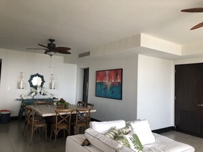 Un salon moderne avec un coin repas, agrémenté d’un ventilateur de plafond, d’un grand tableau abstrait et d’un canapé blanc orné de coussins décoratifs.