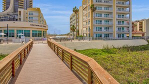 A wooden boardwalk leading to a beachfront hotel with multiple balconies and a restaurant/bar.