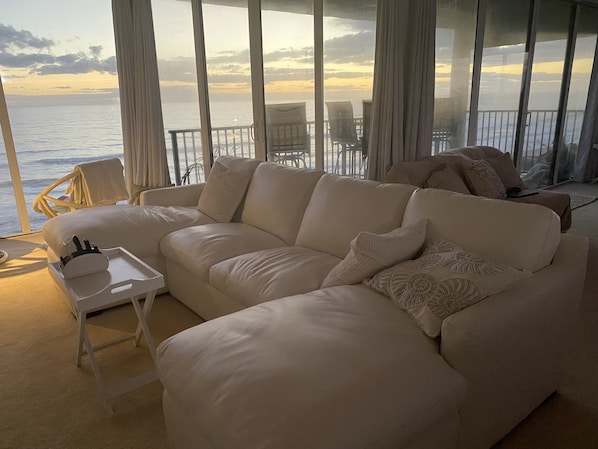 Een woonkamer met een beige bank, een witte bijzettafeltje en uitzicht op de oceaan door grote ramen.