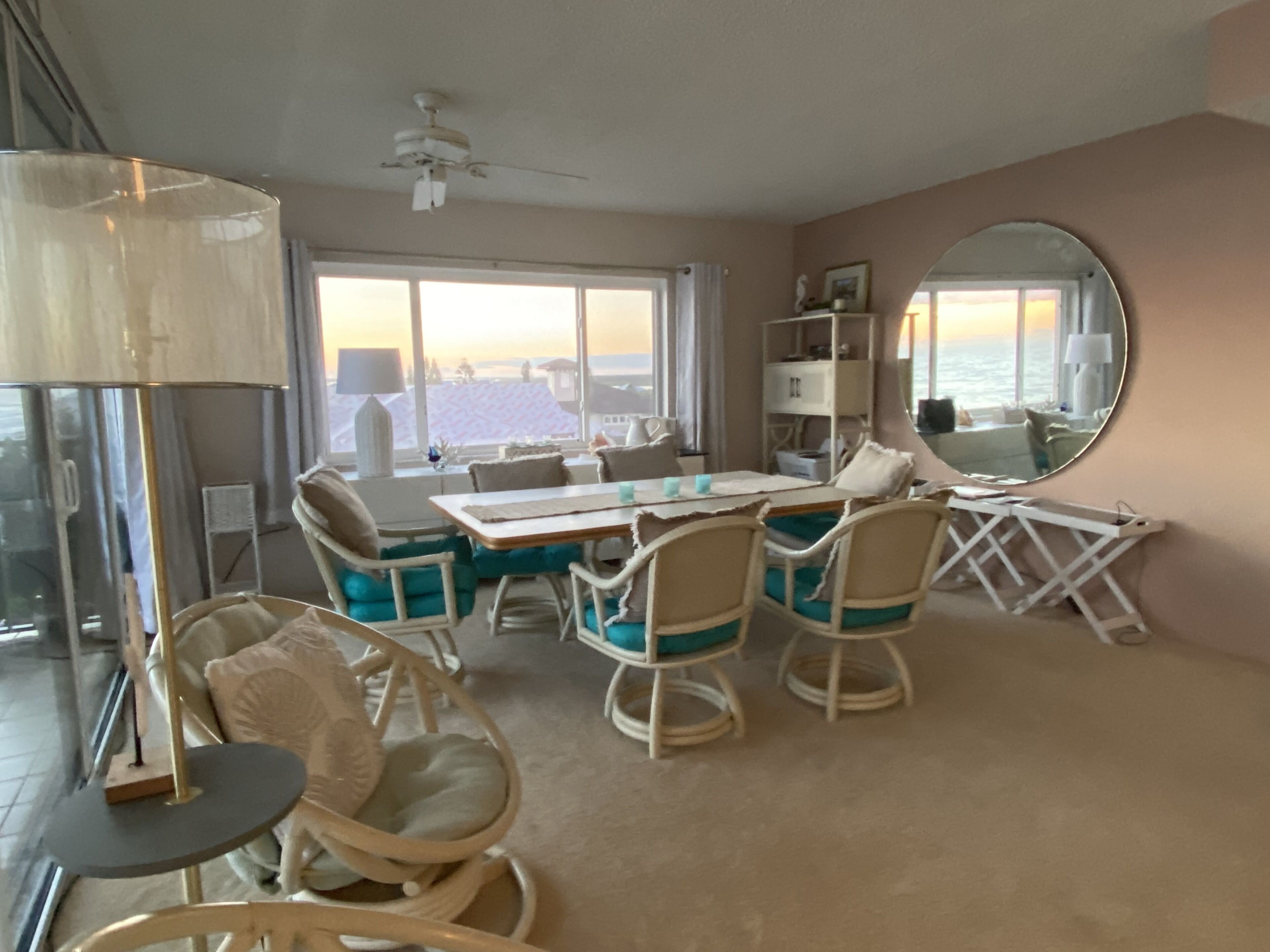Une pièce comprenant une table à manger, des chaises, un ventilateur de plafond, une grande fenêtre avec vue et un grand miroir rond accroché au mur.