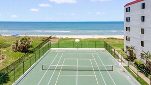 Cancha de tenis exterior con red, rodeada por una valla verde, con vistas a una playa y al océano.