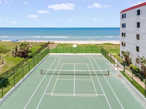 Een tennisbaan buiten met een net, omgeven door een groene hekken, met uitzicht op een strand en de oceaan.