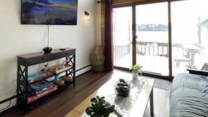 Ett rum med träpanel i tak och på väggarna, ett stort fönster med utsikt över en brygga, en tv på väggen, ett träbord med en växt och en säng.