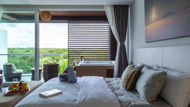 A modern hotel room with a large bed, a bedside table, a window with blinds, a view of greenery, and a painting on the wall.
