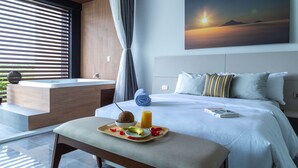 A modern hotel room with a large bed, a view of the sunset, a bathtub, and a breakfast tray with fruit and juice.