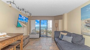 Een balkon met uitzicht op de oceaan en gebouwen, een grijze bank, een houten tafel met stoelen, en een schilderij aan de muur.