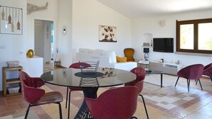 Una sala de estar moderna con una mesa de comedor de vidrio rodeada por sillas rojas, un sofá blanco, un sillón amarillo y un televisor en la pared.