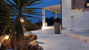 Un patio al aire libre bien iluminado con un sendero de piedra, una palmera y un edificio blanco con ventana y puerta con contraventanas.
