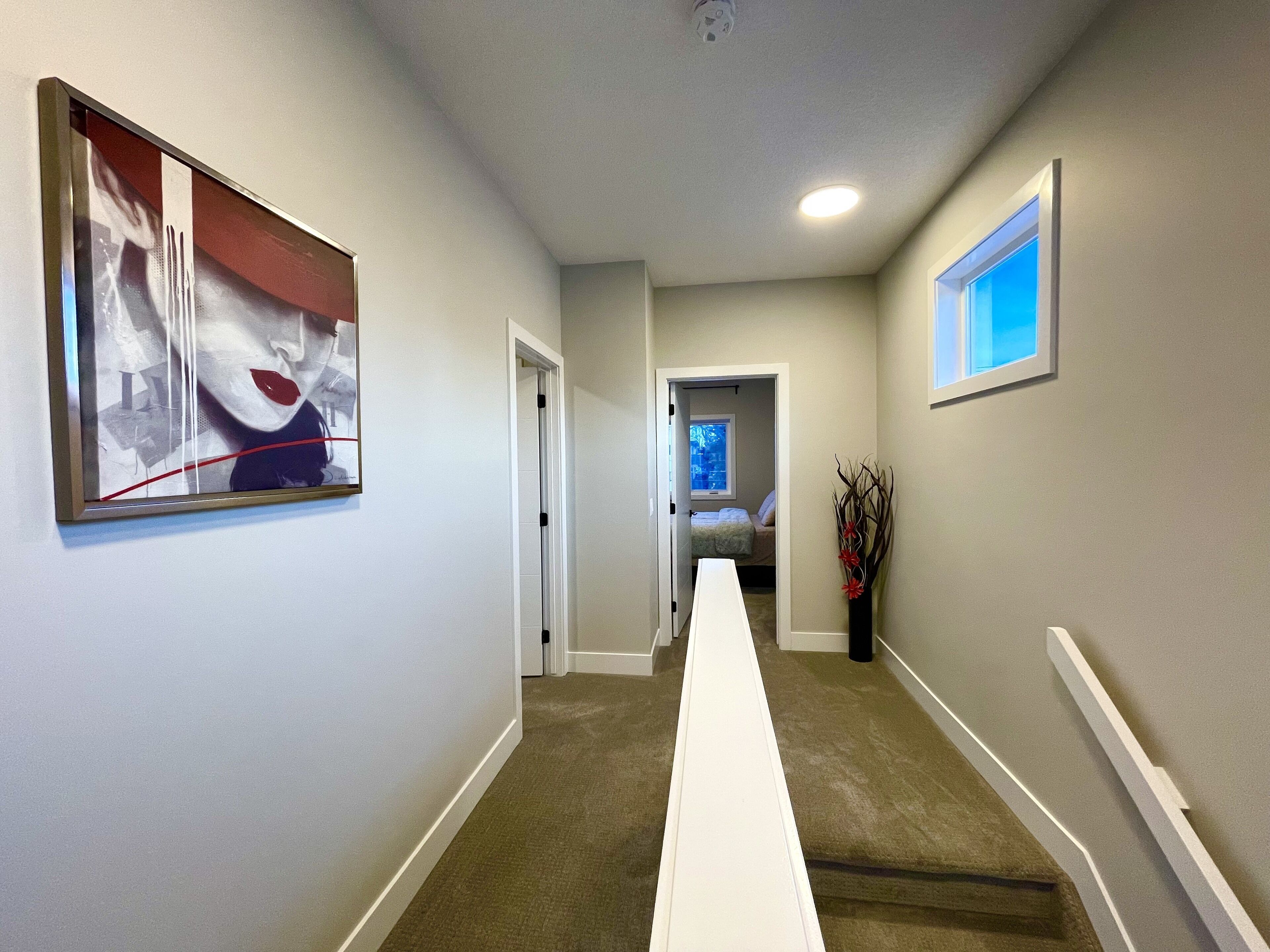 Un couloir avec un long banc blanc, un tableau abstrait encadré, une fenêtre donnant sur un paysage et une porte menant à une chambre.