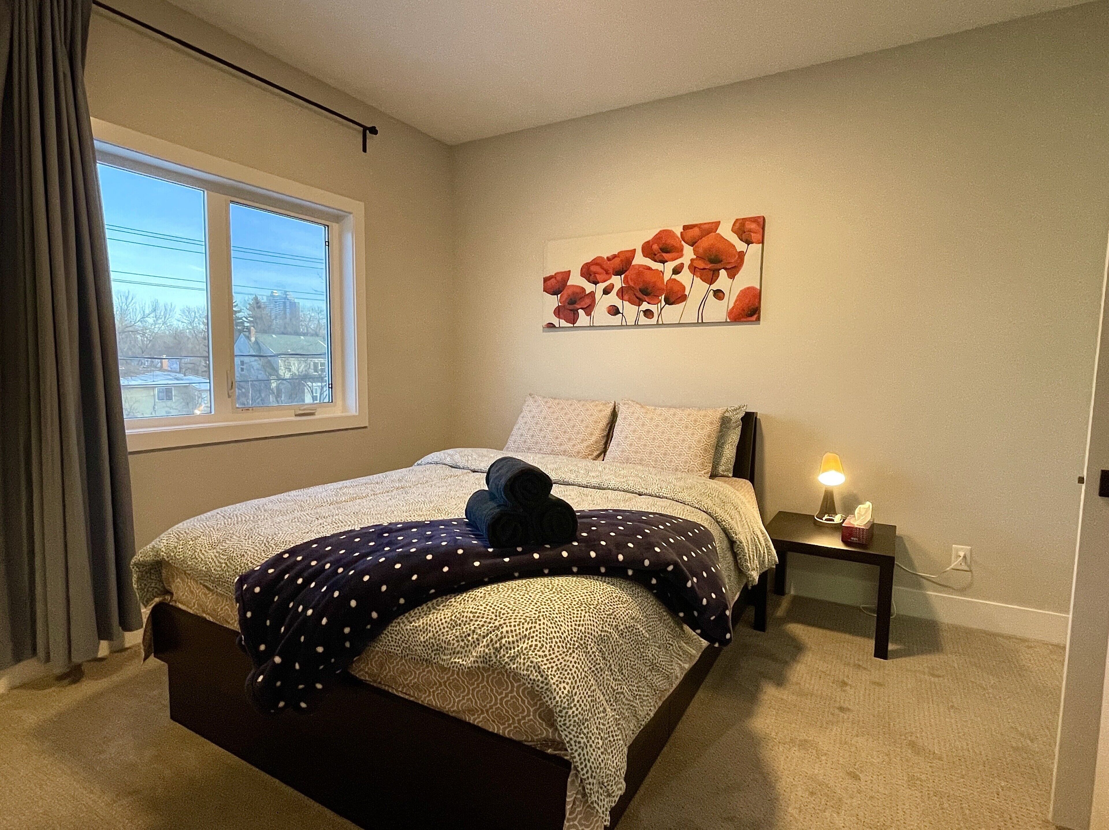 Un dormitorio con cama, ventana, un cuadro y una mesita de noche con lámpara.