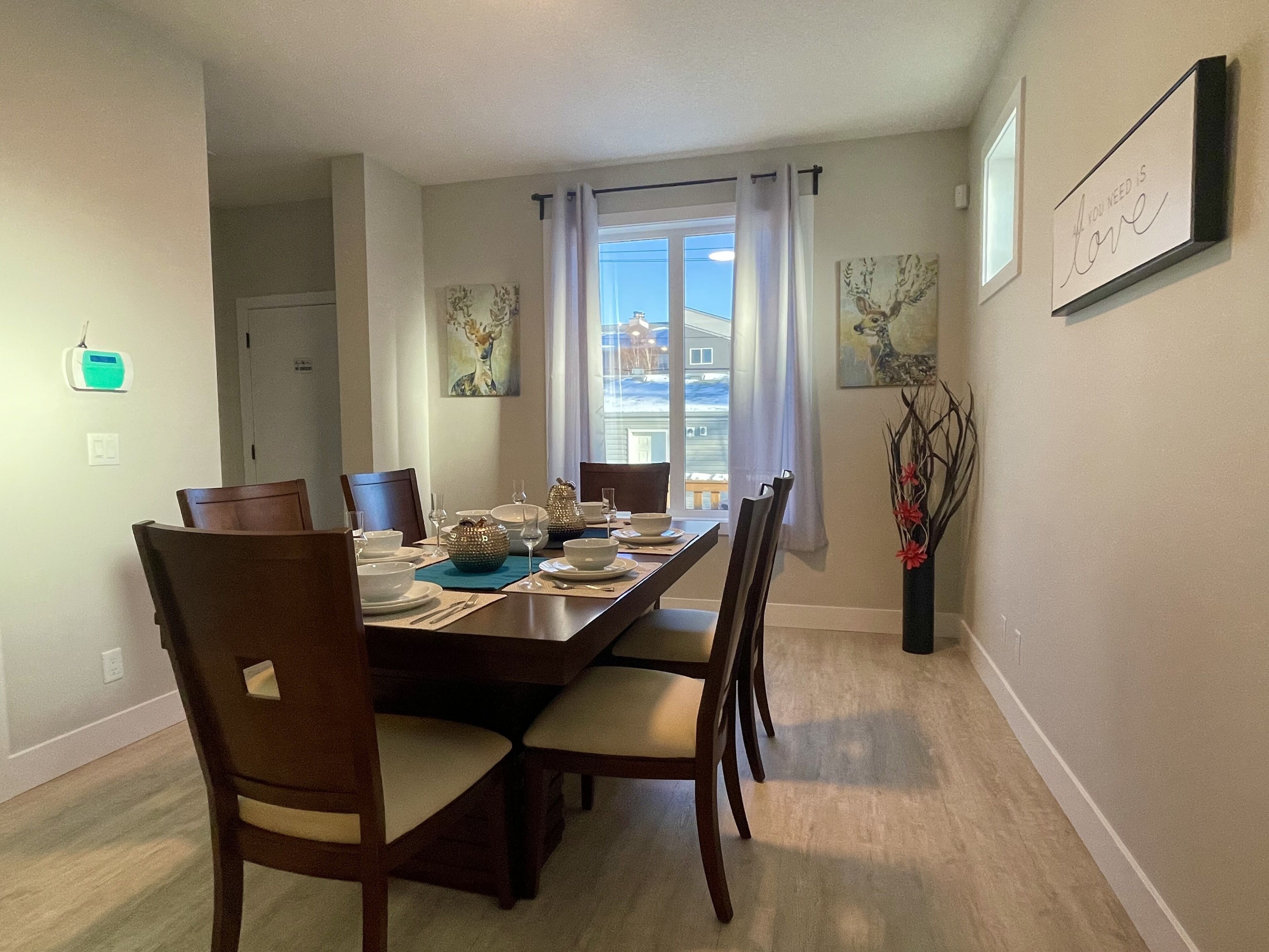 Un coin repas avec une longue table dressée pour quatre personnes, entourée de chaises. Deux tableaux sont accrochés au mur, il y a une fenêtre avec des rideaux et, sur le côté droit, un petit vase contenant des branches.