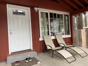A house with a white door, a window with a view of trees, and two lounge chairs outside.