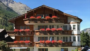 Un hotel a più piani con balconi decorati da fiori rossi, circondato da un giardino curato e vista sulle montagne.