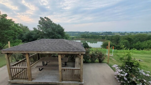 Ein hölzerner Gazebo mit geneigtem Dach und einer Betonplattform, mit Blick auf einen See, umgeben von viel Grün.