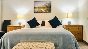 Een hotelkamer met een bed, twee nachtkastjes met lampen, een bankje en een schilderij van een meer en bergen aan de muur.