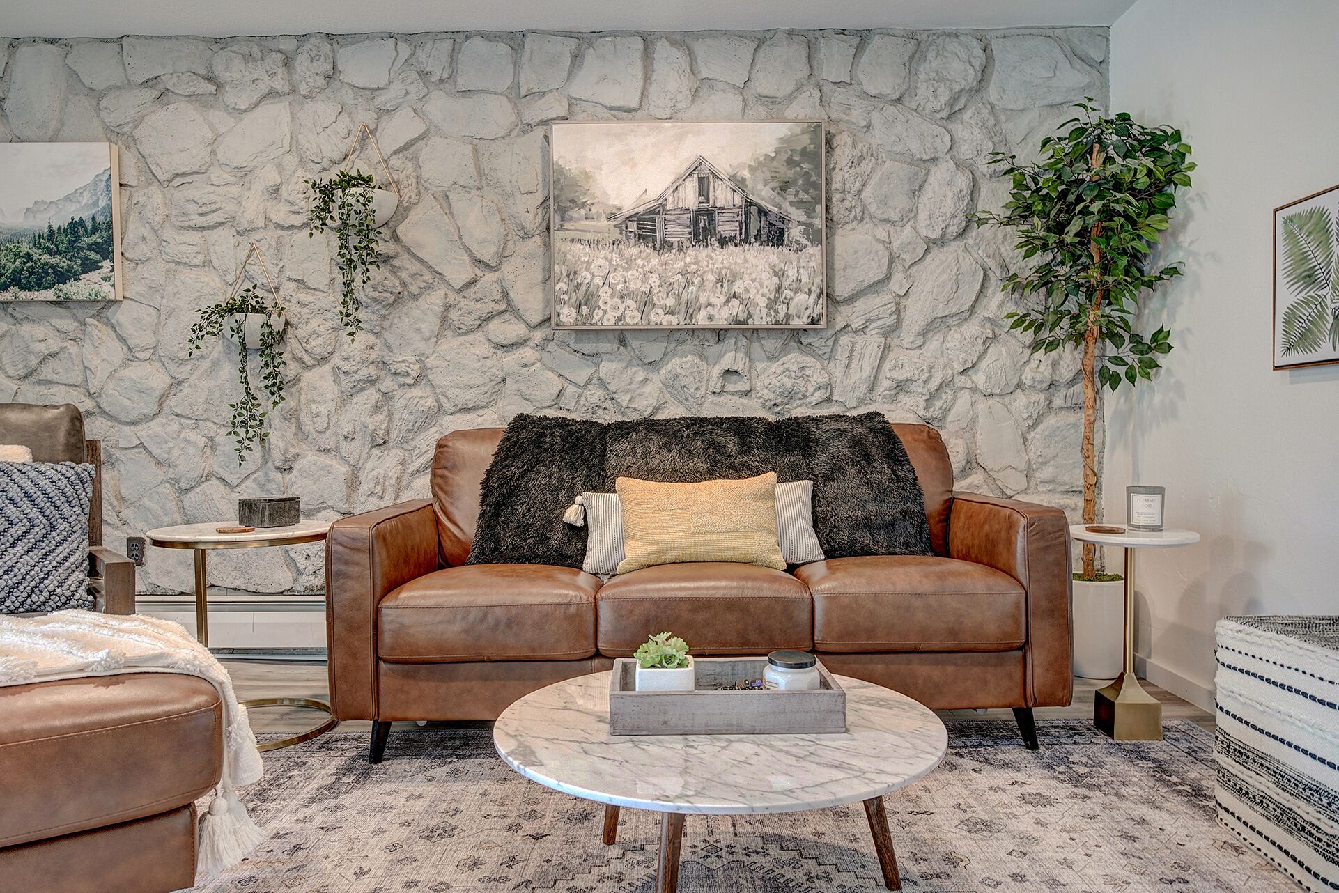 A living room with a stone wall, a brown leather sofa, a round coffee table, and a painting of a barn.
