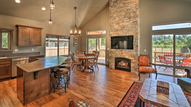A spacious kitchen with a large island, wooden dining table, stone fireplace, and large windows offering views of the outdoors. - Rustic Cabin Near Mt Rainier: Fire Pit & BBQ! (Packwood)