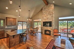 A spacious kitchen with a large island, wooden dining table, stone fireplace, and large windows offering views of the outdoors. - Rustic Cabin Near Mt Rainier: Fire Pit & BBQ! (Packwood)