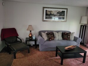 A living room with a grey sofa, patterned cushions, a black armchair with a red throw, a side table with a lamp, a framed landscape painting, and a small black table with a decorative bowl.