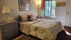 Un dormitorio con cama, mesita de noche, lámpara, ventana con cortinas, puerta y pared decorada con una bandera estadounidense enmarcada y un letrero motivacional.