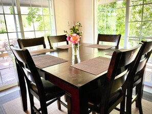 A dining table with six chairs, a vase with flowers, and a view of greenery through large windows.