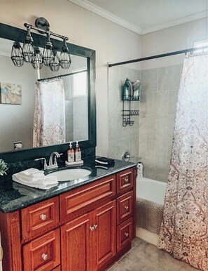 Un baño con tocador de madera, un espejo grande, ducha con cortina estampada y un perchero para toallas.