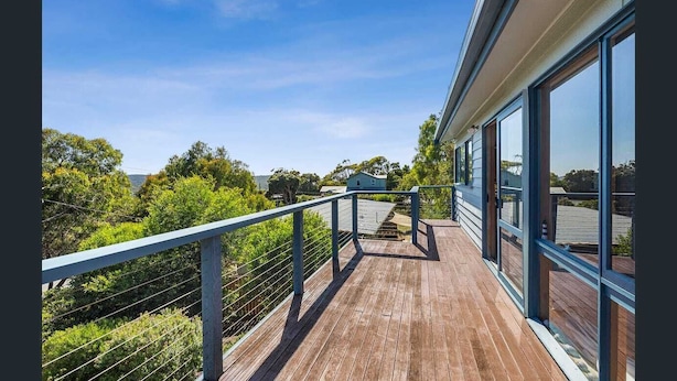 A wooden deck with a metal railing, large windows, and a view of greenery and a distant building. - BEAUTIFUL BEACH HOUSE - BRILLIANT POSITION (Anglesea)