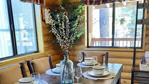 A wooden dining table set with white plates, bowls, and wine glasses, flanked by cushioned chairs. A vase with green foliage is placed on the table.