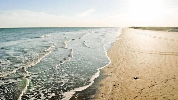 A beach with waves reaching the shore, a clear sky, and a sandy path leading along the water.