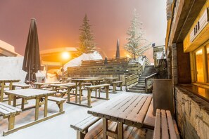 Des bancs et des tables en bois, installés à l’extérieur, sont recouverts de neige. On aperçoit, en arrière-plan, un mur de pierre et un bâtiment en bois.