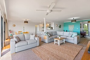 Ein modernes Wohnzimmer mit einer grauen Sofagarnitur, einem Couchtisch und einem großen Teppich. Ein Deckenventilator ist vorhanden, im Hintergrund ist eine Küchenzeile zu sehen, und eine Wand ist in Türkis gehalten.