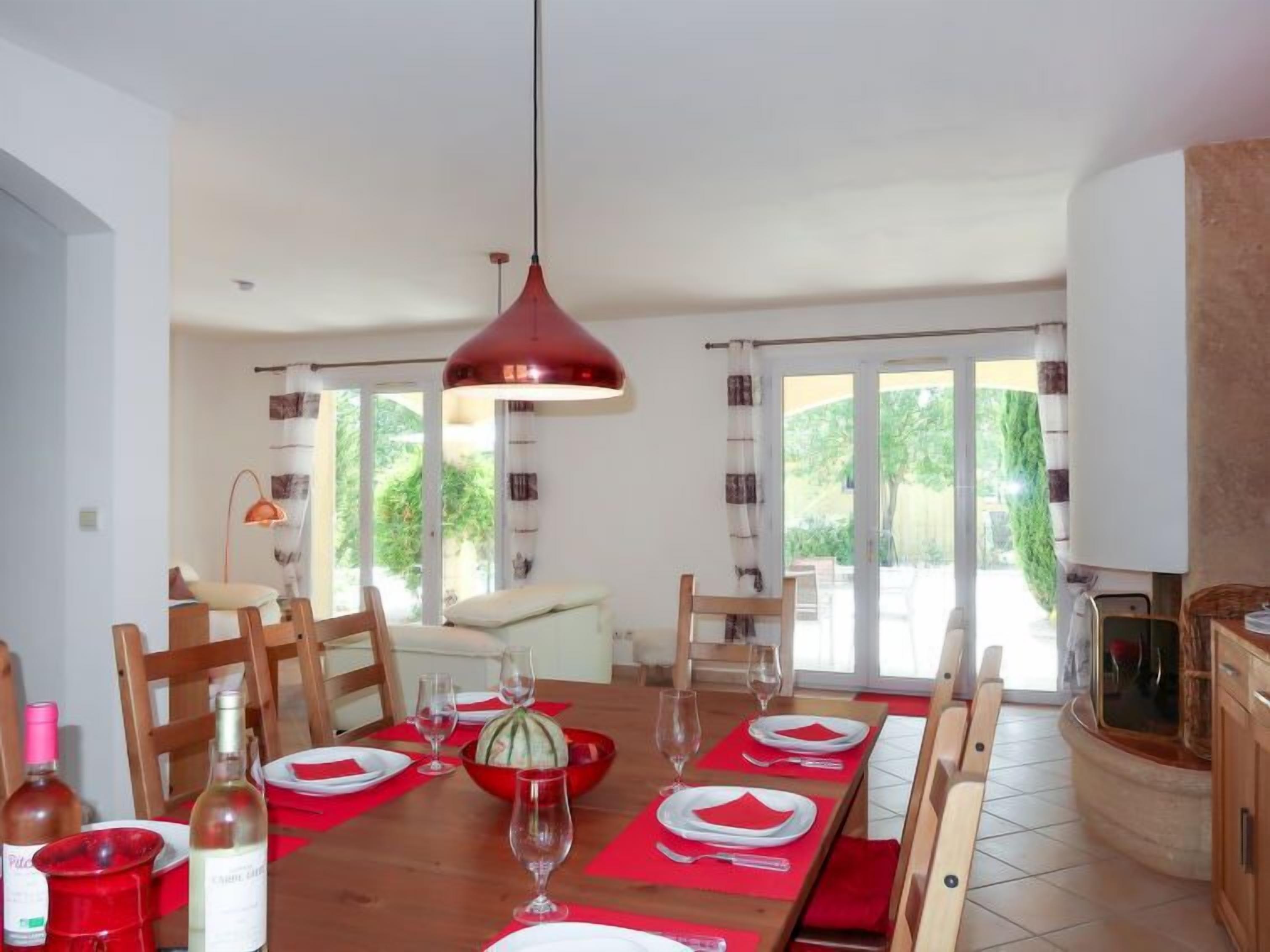 Un coin repas avec une longue table en bois dressée pour le repas, des sets de table rouges et un luminaire suspendu rouge.