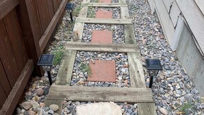 Ein schmaler Pfad aus Holzplanken und Steinplatten, begrenzt von einem Holzzaun und einem Haus.