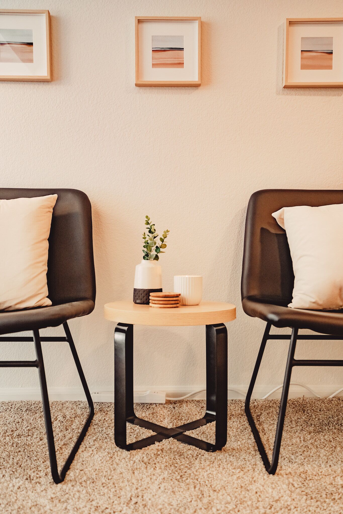 A room with two black leather chairs, a small round table with a plant and a white vase, and framed pictures on the wall.