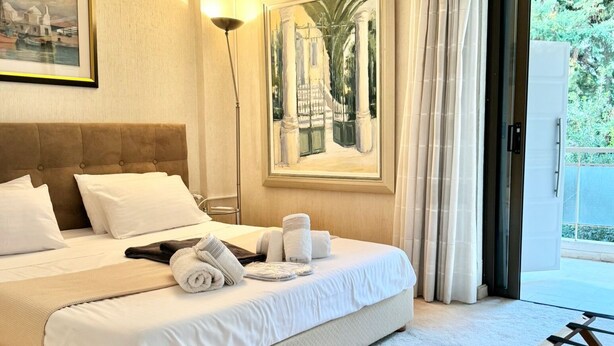 A hotel room with a large bed, a painting of a building, and a view of palm trees through the window.