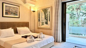 A hotel room with a bed, pillows, towels, a painting, a lamp, and a view of trees through a window.